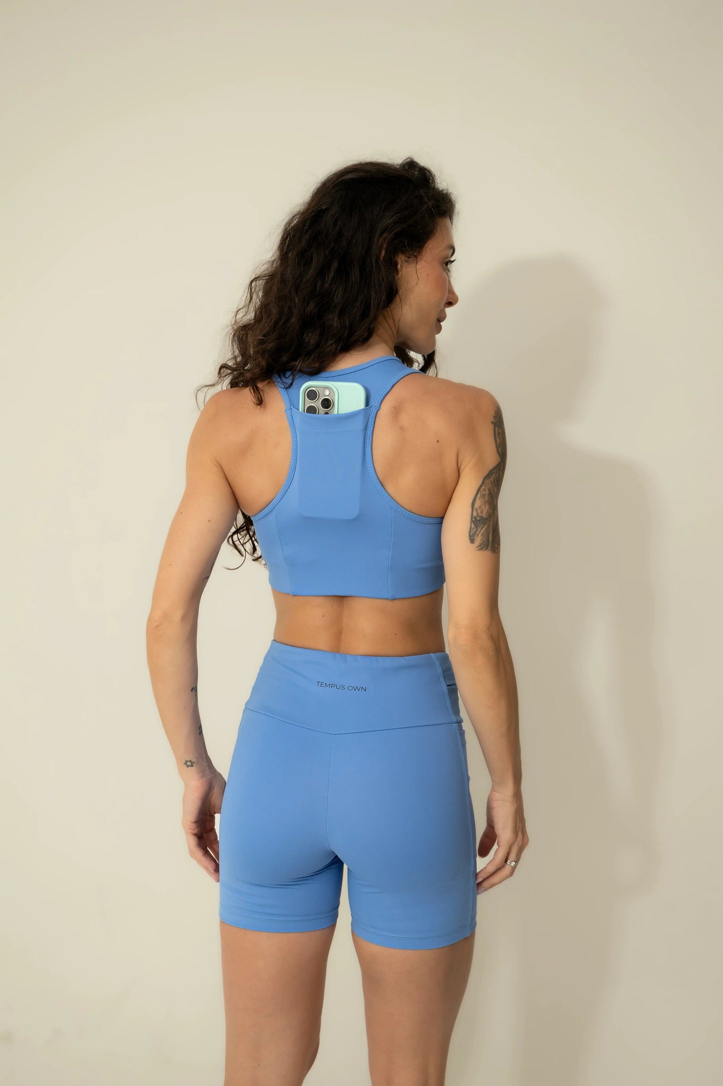 Back view of a woman running in a Caravela Blue racerback sports bra, demonstrating the integrated back pocket for secure phone storage
