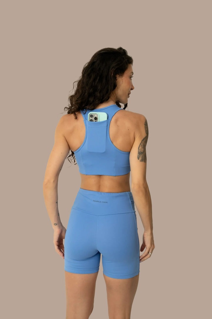 Back view of a woman running in a Caravela Blue racerback sports bra, demonstrating the integrated back pocket for secure phone storage