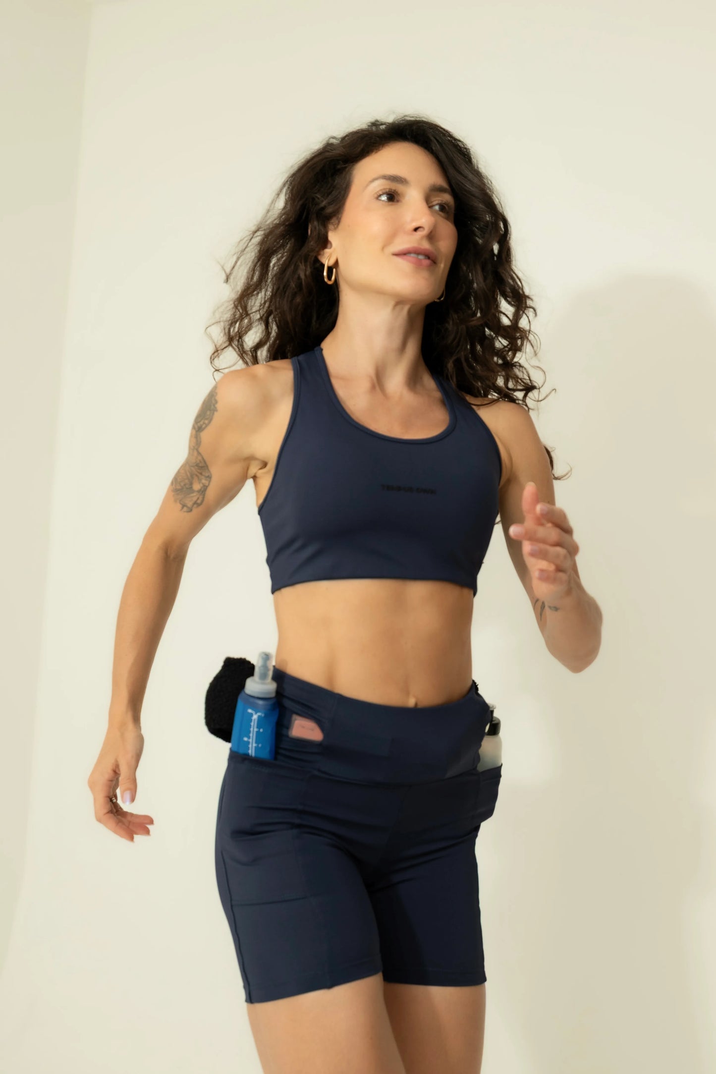Action shot of a woman running in Deep Blue high-waisted shorts, showing how the secure pockets hold a water bottle and nutrition firmly during exercise.