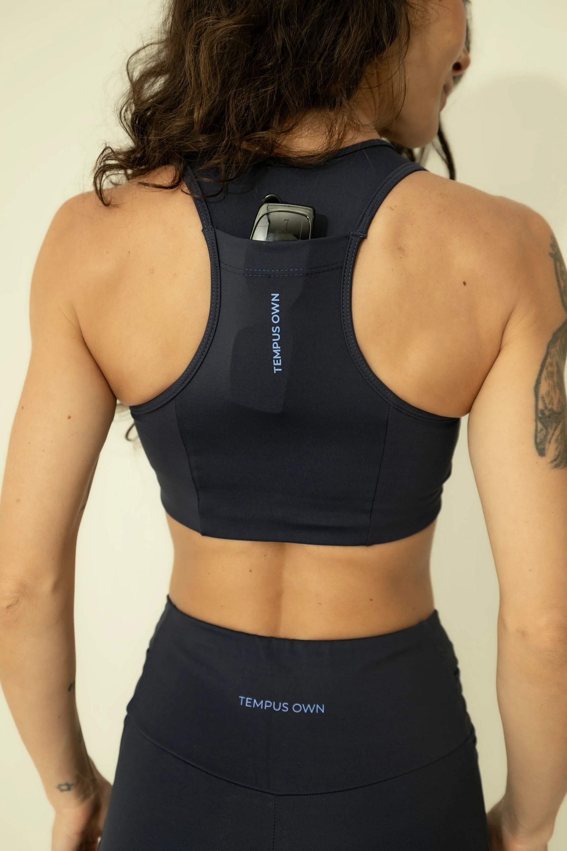 Back view of the Deep Blue racerback sports bra, demonstrating the functional back pocket being used for secure sunglasses storage during outdoor training.