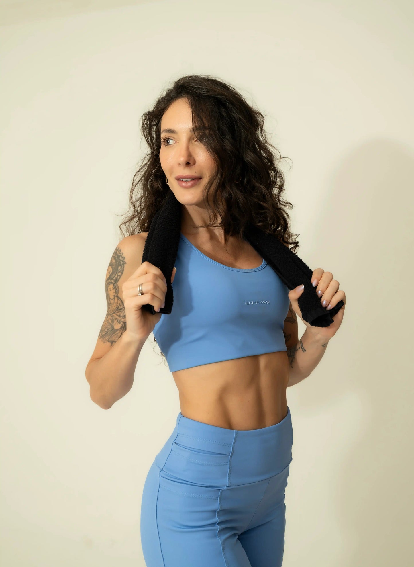 Lifestyle studio shot of a woman holding a towel after a workout, wearing the Caravela Blue high-support sports bra by Tempus Own, showcasing the flattering front fit and premium athletic design.